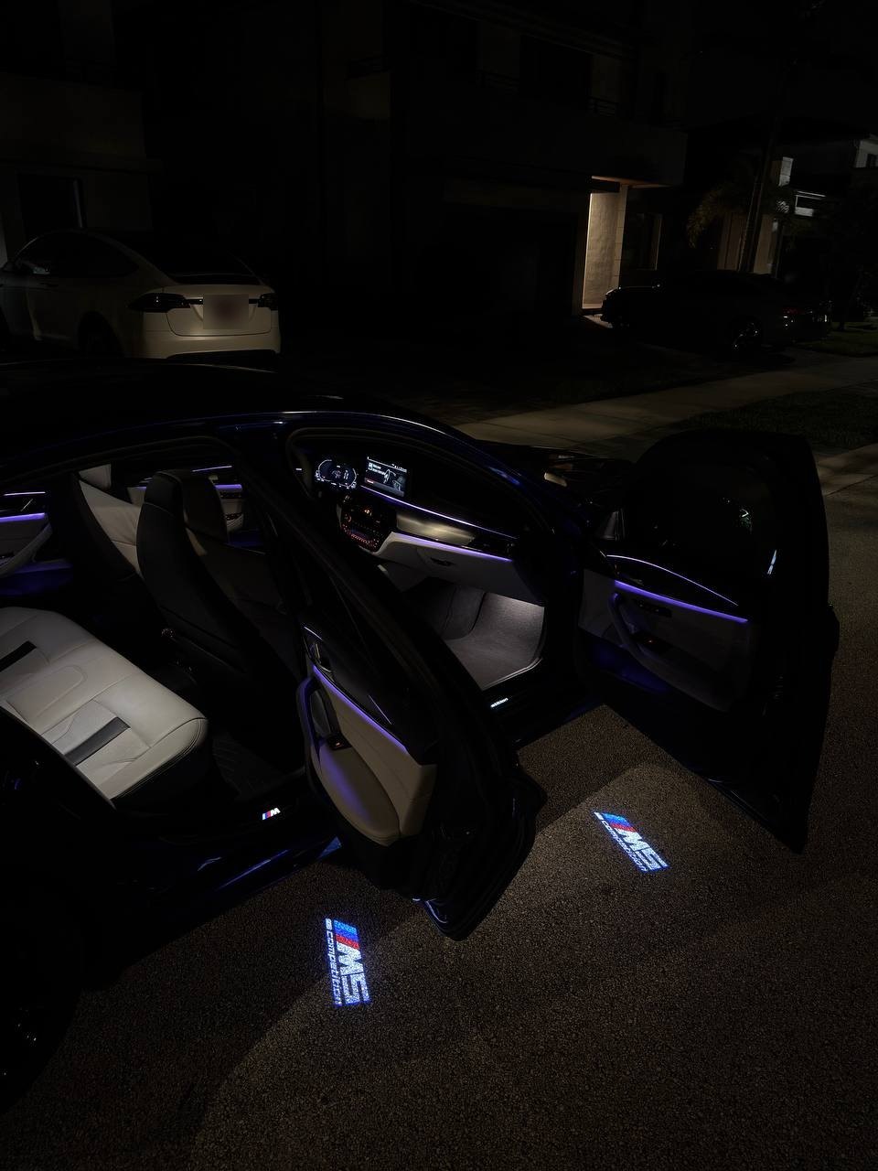 Front and rear doors open on a BMW M5 at night with door lights projecting M5 logos