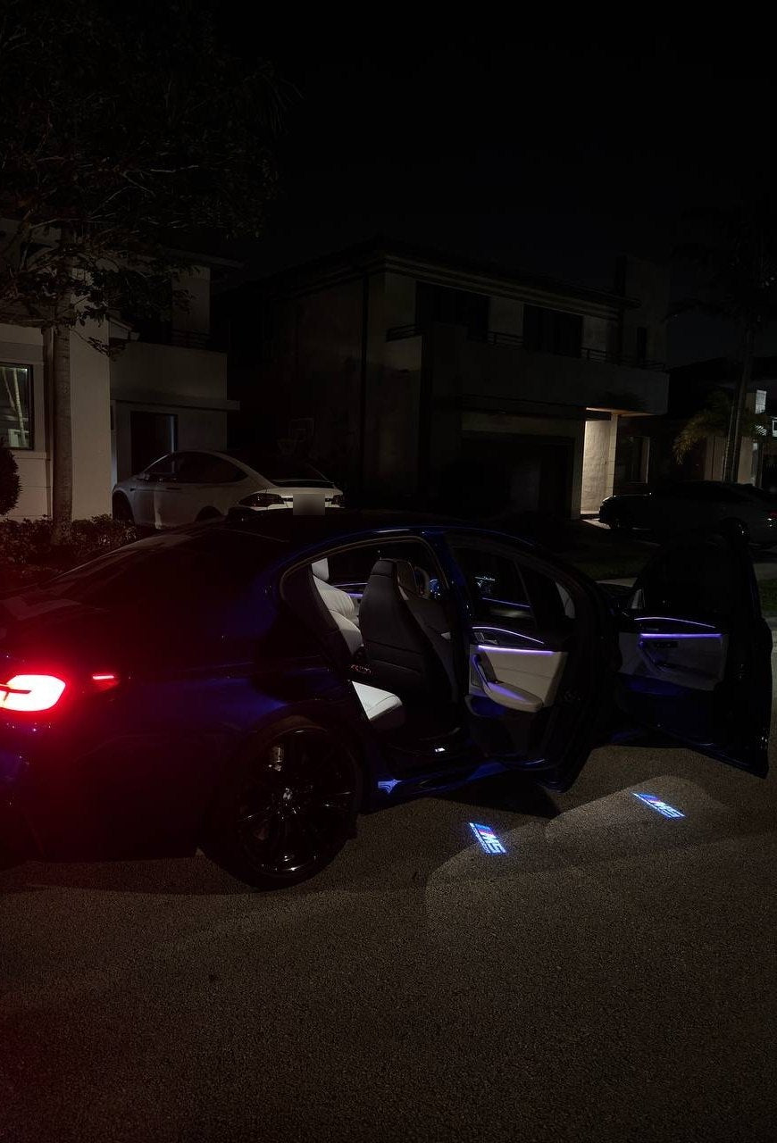 Side view of a BMW M5 at night, doors open, door lights projecting logos on the street